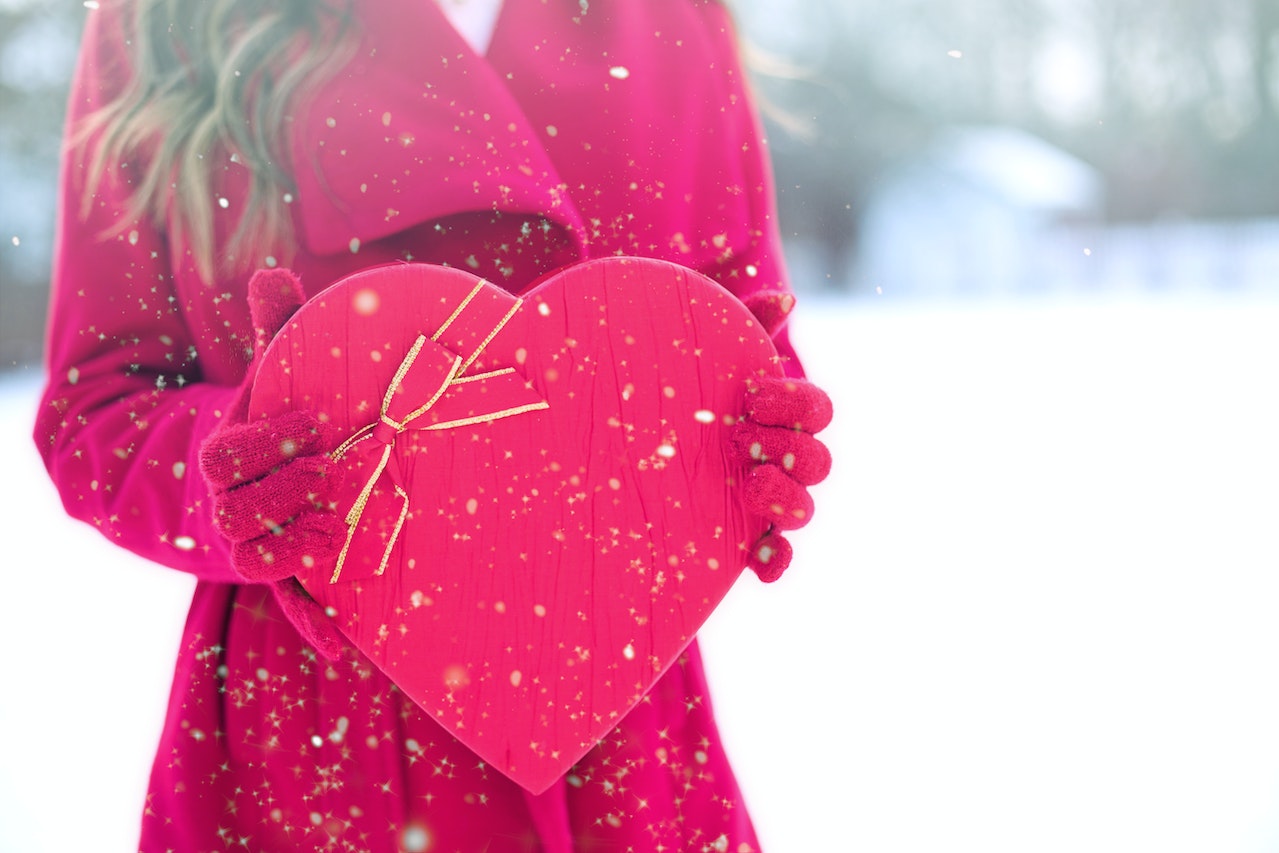 A person wearing a red winter coat while wearing red gloves and holding a red heart-shaped package