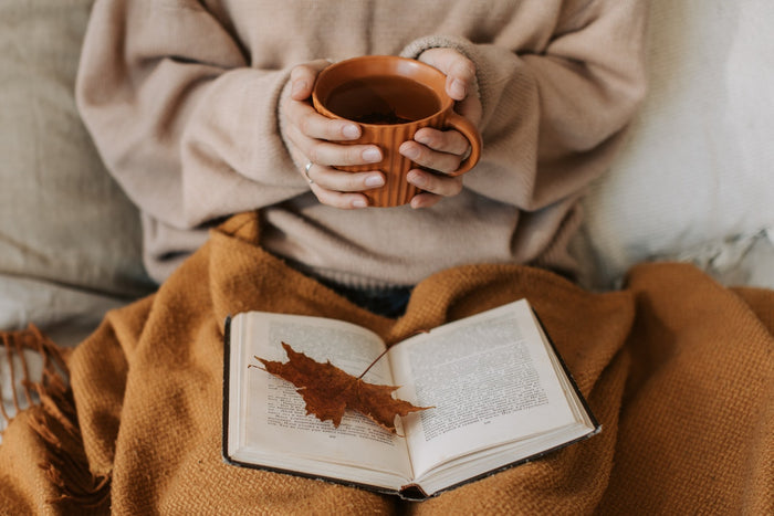 A person in a light brown sweater sitting on a bed while holding a cup of tea and reading a book on her lap